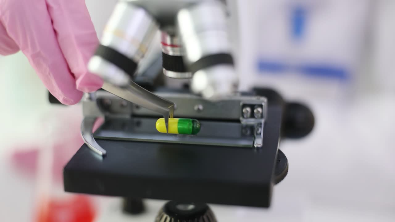 A hand in a pink glove places a medicine capsule under a microscope in a laboratory
