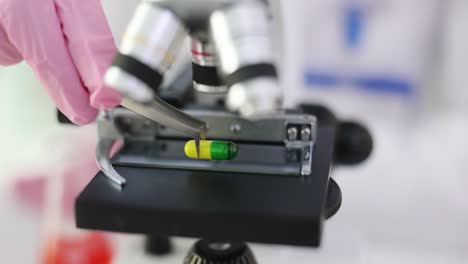 a hand in a pink glove places a medicine capsule under a microscope in a laboratory