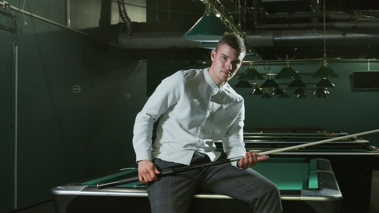 молодой человек в белой рубашке и серых брюках сидит на зеленом бильярдном столе с легкой улыбкой, держа в руках палку. навесные лампы отбрасывают мягкое освещение, современный интерьер на заднем плане, видны несколько бильярдных столов