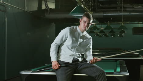 un joven con camisa blanca y pantalones grises se sienta en una mesa de billar verde con una sonrisa ligera, sosteniendo un palo de taco. las lámparas aéreas emiten una iluminación suave, un interior moderno en el fondo, varias mesas de billar visibles