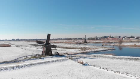 Patinadores-Sobre-Hielo-En-El-Canal-Congelado-Al-Lado-Del-Molino-De-Viento-De-Holanda,-Vista-Aérea-De-Invierno