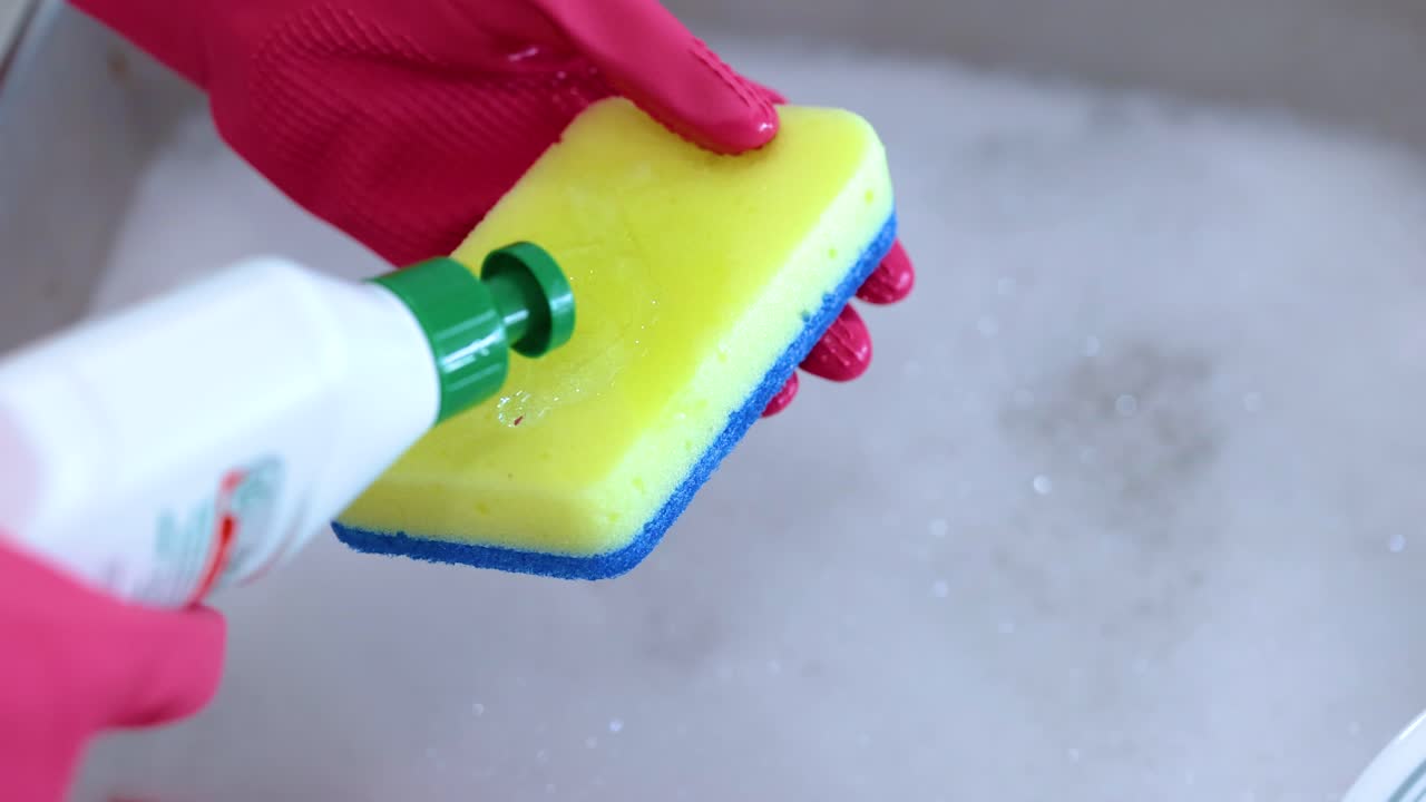 A person wearing red gloves applies dish soap to a yellow sponge over a sink filled with soapy water