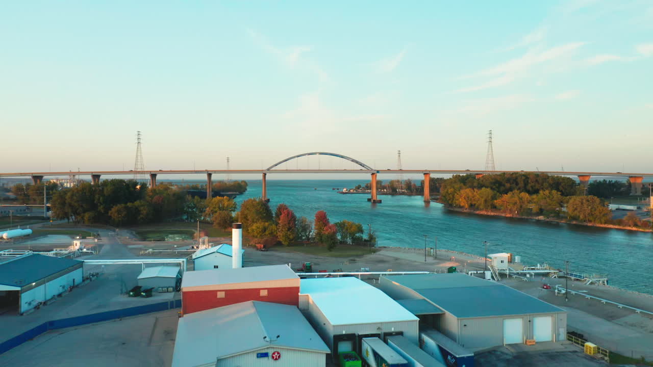 The graceful blue arch of the Leo Frigo Bridge stretches across a broad river framed by autumn trees and industrial docks, glowing gently in the fading dusk light