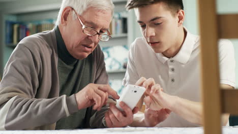 Junge-Pflegekraft,-Die-Mit-Einem-älteren-Mann-Mit-Brille-Spricht-Und-Ihm-Dabei-Hilft,-Die-Smartphone-Anwendung-Zu-Verwenden