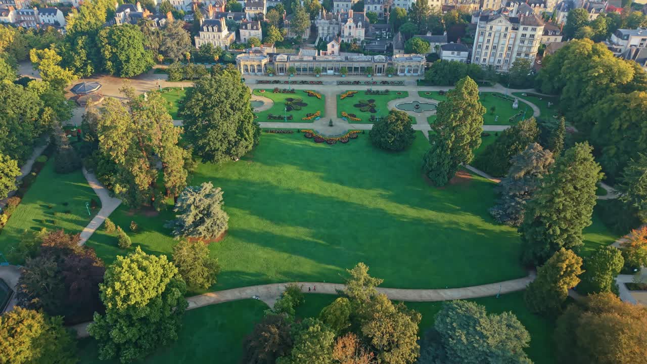 Drone pulls back over Thabor Park in Rennes, showing the bandstand, fountains, paths, French-style garden, surrounding buildings and sunrise light