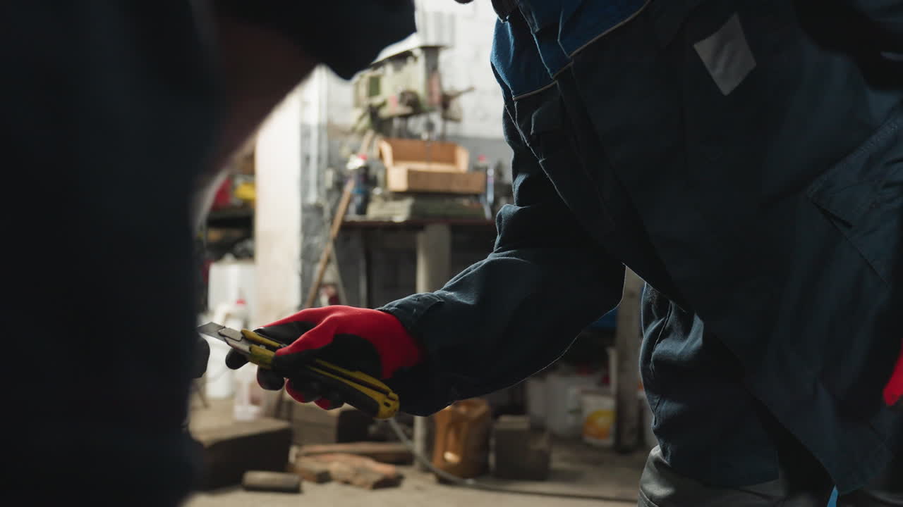Mechanic in blue uniform wearing red gloves holds a yellow scraper while working in an automotive workshop, background features tools, equipment, and mechanical parts