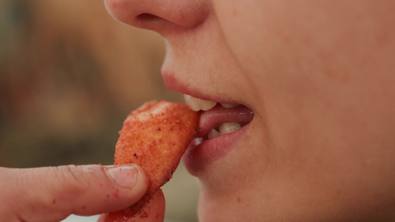 Vista de perfil fechada de mulher adulta provando um lanche de laranja, a língua tocando a comida perto dos lábios entreabertos, os dedos segurando-o, a mandíbula fechando em movimento natural de alimentação com características faciais detalhadas e luz suave