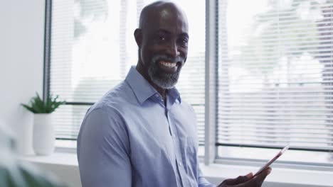 Smiling-african-american-businessman-using-digital-tablet-and-standing-by-window-in-office