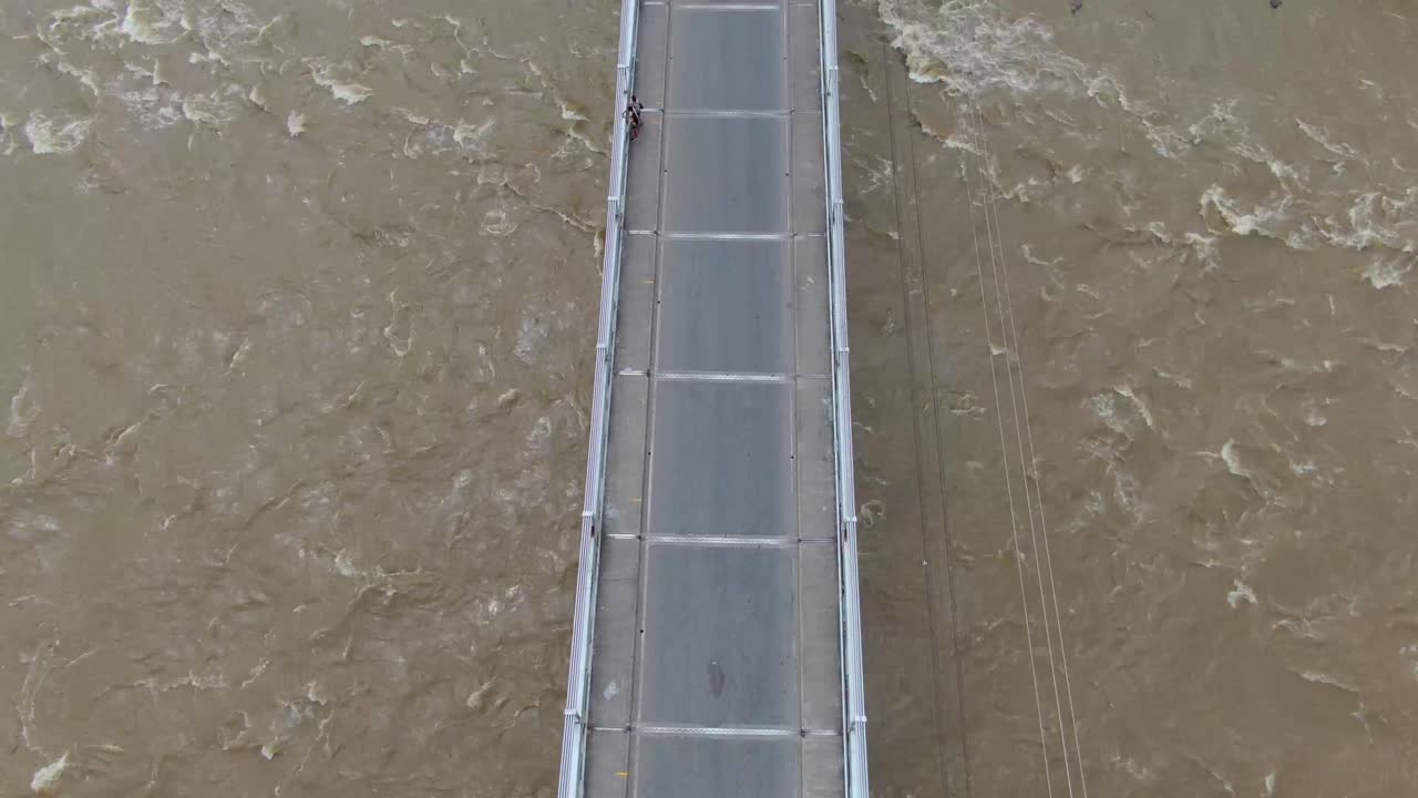 Top down view of suspension bridge with rushing brown river and jagged rocks