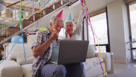 Pareja-Caucásica-Mayor-Soplando-Sopladores-De-Fiesta-Celebrando-Un-Cumpleaños-Mientras-Realiza-Una-Videollamada-En-Lapt