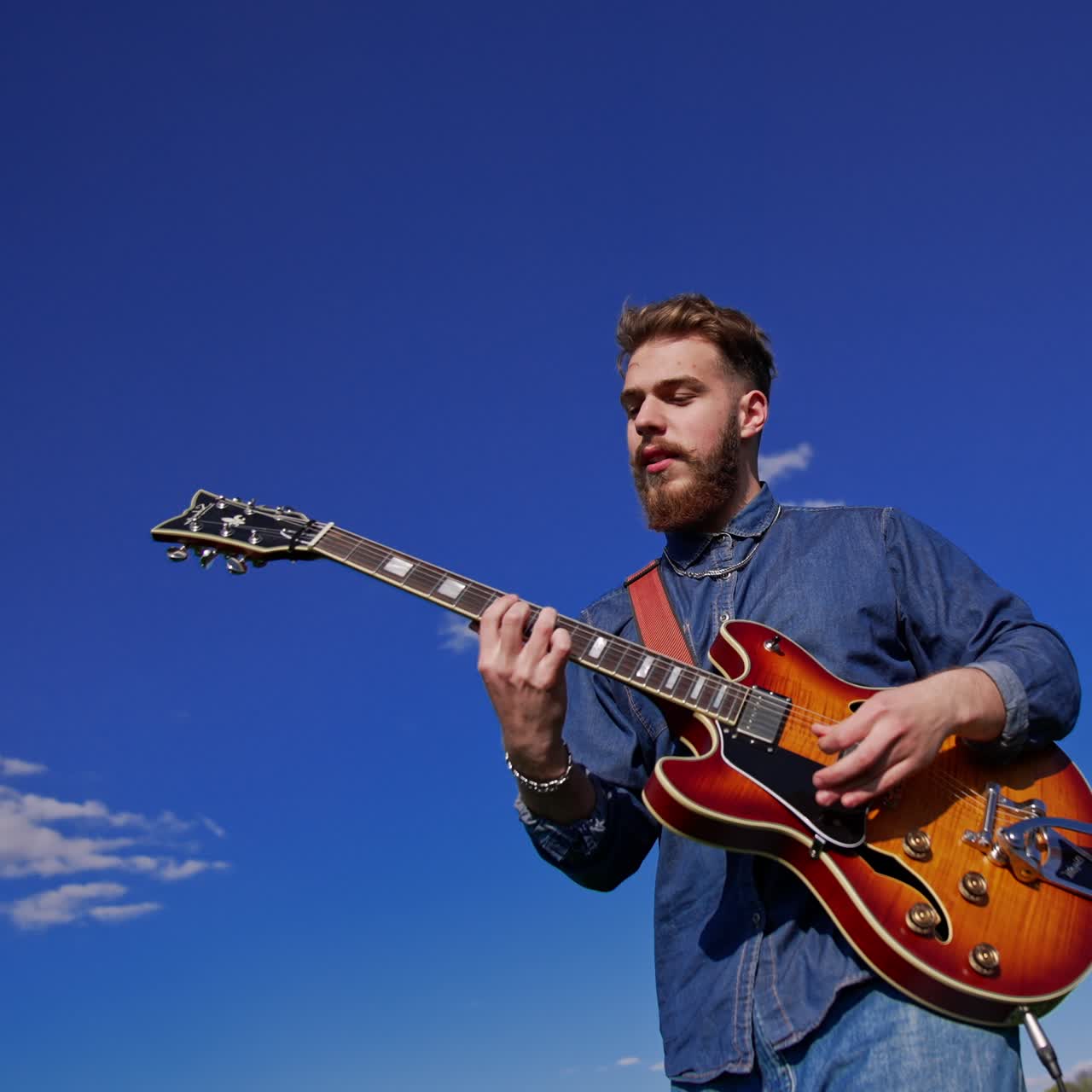 Bärtiger, junger Mann im Jeanshemd spielt energiegeladen Gitarre und singt. Gitarrist tritt im Freien vor dem Hintergrund des blauen Himmels an einem sonnigen Tag auf.