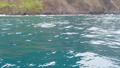 Hd-120fps-Hawaii-Kauai-Bootfahren-Auf-Dem-Ozean-Delfine-Plätschern-In-Der-Ferne-Und-Ein-Delfin-Bläst-Wasser-Mit-Felsigen-Klippenhintergrund