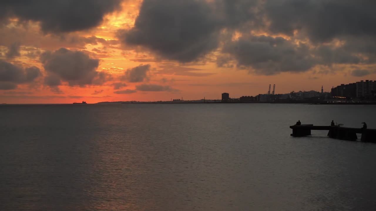 Kaunis kultainen auringonlasku heittää heijastuksia tyynelle merelle. Kaupungin siluetti näkyy etäällä siluetteineen laiturilla.