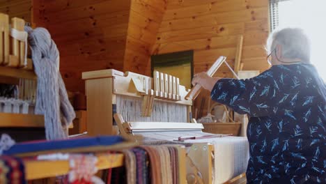 Side-view-of-old-caucasian-senior-woman-weaving-cloth-on-handloom-machine-in-a-workshop-4k