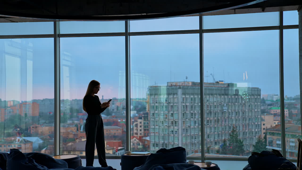Full-length portrait on a business woman in modern office. Panoramic view on large office windows with cityscape indoors. Beautiful female business worker with a phone.