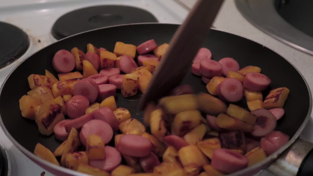 person mixing potatoes and sausages in a frying pan on electric stove with a wooden spatula, close-up