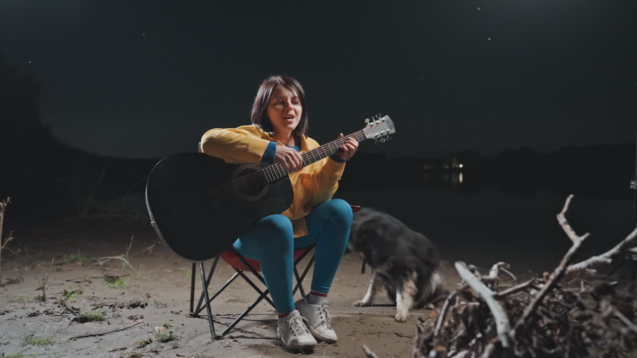 Guitarspil på stranden om natten. En kvinde spiller guitar ved lejrbålet. En asiatisk kvinde spiller guitar ved siden af sin hund under stjernerne. En hyggelig aften ved kysten, hvor hun spiller guitar ved siden af sin loyale hund under en stjerneklar himmel.