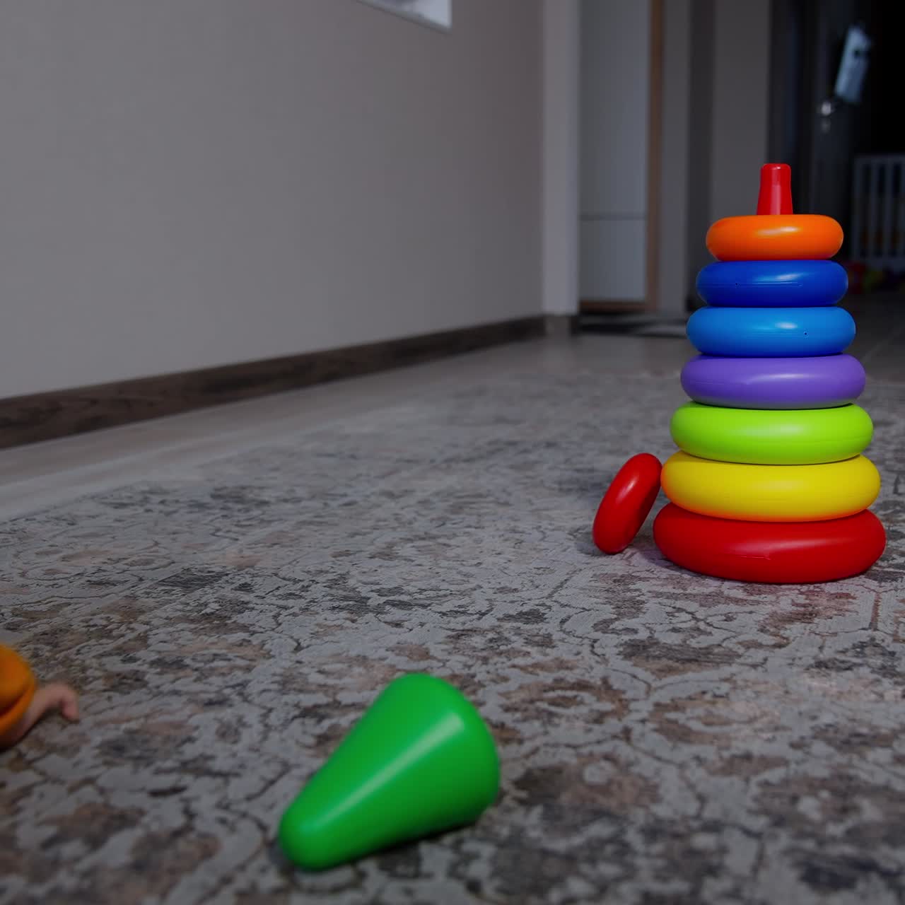Baby crawling along the room ignoring bright toy pyramid. Boy in orange sport suit stands up holding by the wall
