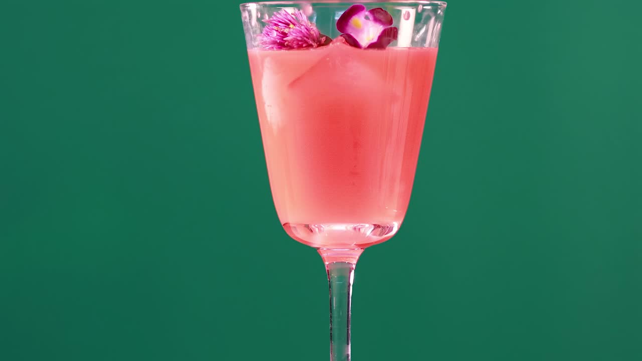 A vibrant pink drink with ice and flower garnish in a glass against a green backdrop.