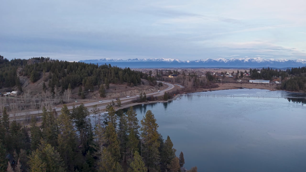 Aerial drone view of a forest-lined road curving along a reflective river or lake with distant snowcapped mountains under a blue sky