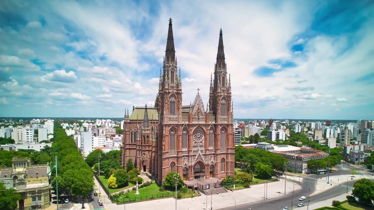 Ripresa aerea in tracking della Cattedrale di La Plata durante una giornata luminosa. Orbita cinematografica dall'alto che mostra l'architettura neogotica, le guglie e la disposizione urbana della città sotto un cielo blu.