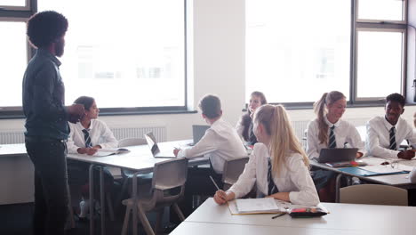 Male-High-School-Teacher-Standing-In-Classroom-Teaching-Lesson-To-Pupils-Wearing-Uniform