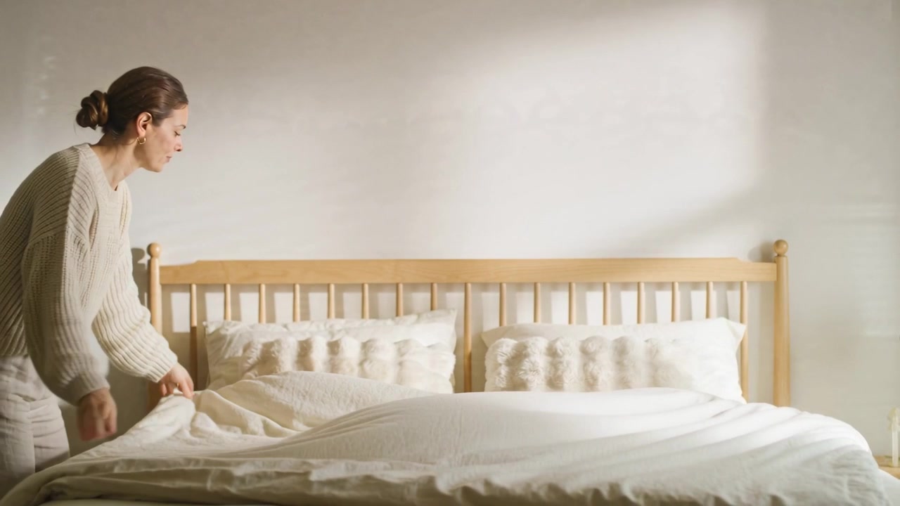 Sunlight urging lady in cream lifting duvet, smoothing pillows in bedroom by headboard, copy space