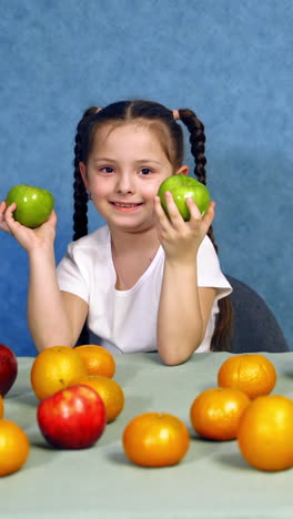 portrait of a cute little girl with fruits indoors. ripe fruit on the table. beautiful smiling child playing with fresh apples on the table. vertical video