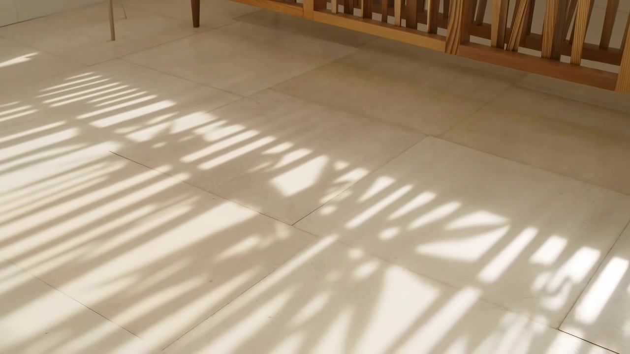 Varmt dagslys falder ind over et rent beige flisegulv i et stille rum, hvilket skaber markante stribede skygger fra et nærliggende vindue eller gelænder og en rolig minimalistisk atmosfære.