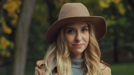 vista de cerca de una joven rubia caucásica con un sombrero sonriendo y mirando la cámara en el parque en otoño