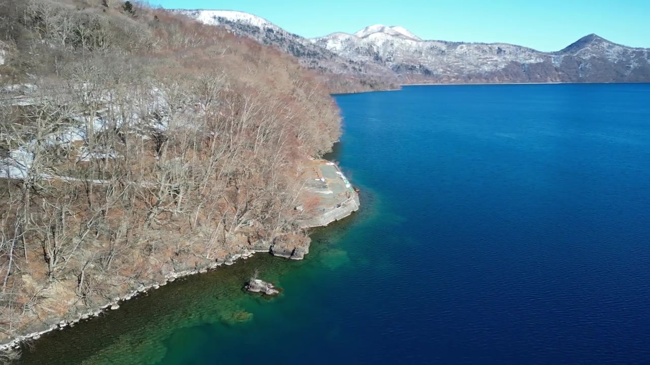 Çıplak kış ağaçları ve uzaktaki karlı dağlarla çevrili kayalık bir kıyı şeridinin yanındaki canlı mavi göl suyunun üzerinde süzülen havadan çekim.