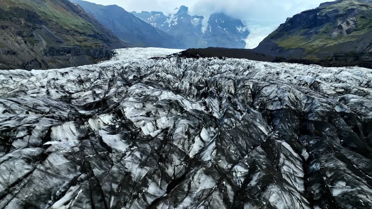 A drone glides above a rugged Icelandic glacier filled with dark volcanic streaks, revealing deep crevasses and distant Nordic mountains under cold, dramatic light