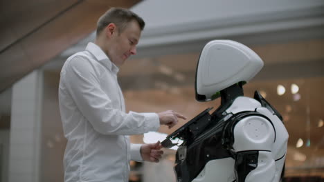 un hombre se para con un robot bot y le hace preguntas y pide ayuda haciendo clic en la pantalla en el cuerpo del robot. la interacción humano-robot en el mundo moderno