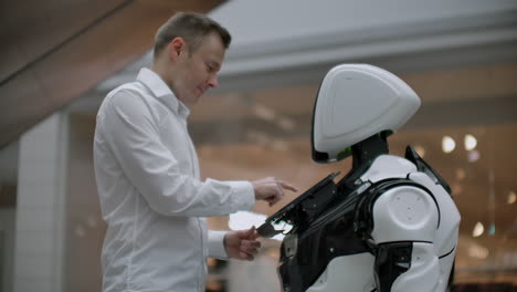 a man stands with a robot bot and asks him questions and asks for help by clicking on the screen on the robot body. human-robot interaction in the modern world