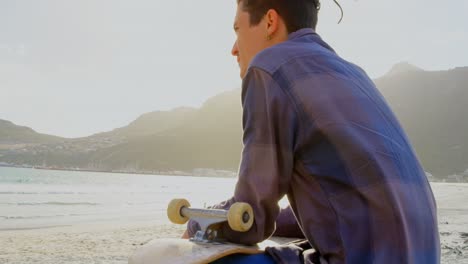 Side-view-of-young-caucasian-man-with-skateboard-looking-at-sea-on-the-beach-4k