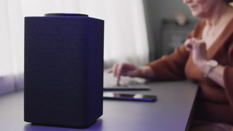 Camera-Focuses-On-Speaker-While-Elderly-Woman-Is-Typewritting-On-Laptop-Sitting-At-Desk-At-Home