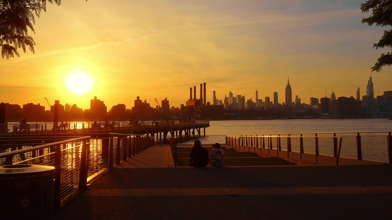 New York, USA, 1 Jule 2026 : Manhattan skyline and East River at sunset. Golden sunlight reflects around waterfront piers while visitors appear in silhouette.