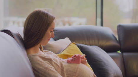 Schwangere-Frau-Sitzt-Entspannt-Zu-Hause-Auf-Dem-Sofa-Und-Schaut-Sich-Kleine-Babyschuhe-An