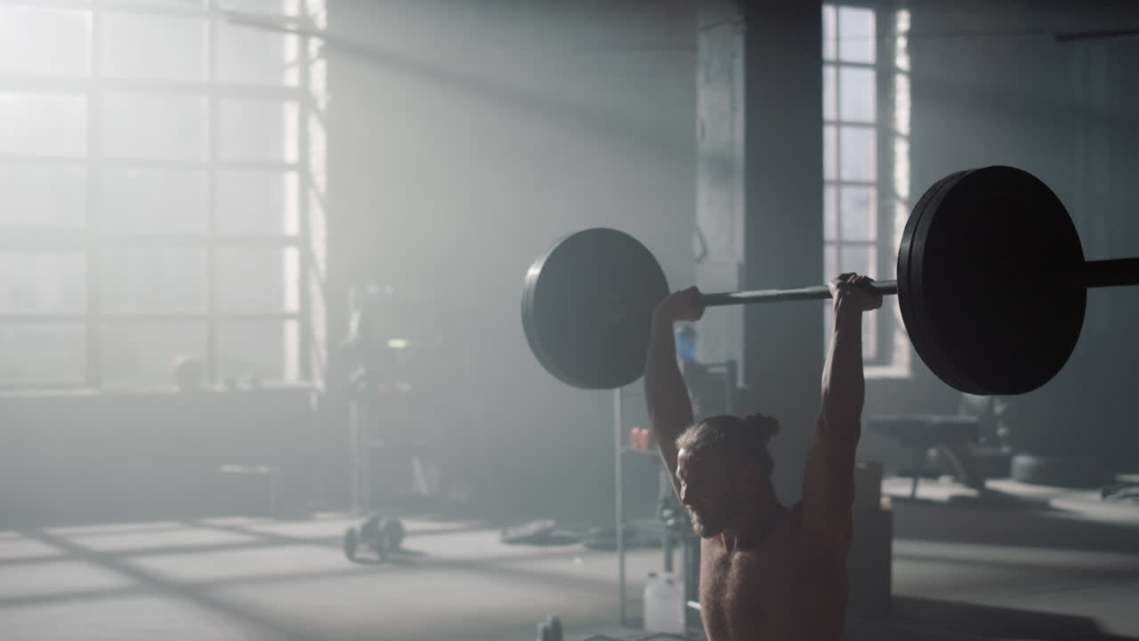 강한 스포츠맨이 무게를 들어 올리는 동안 바벨로 오버헤드 스 ⁇ 을 하는 남자