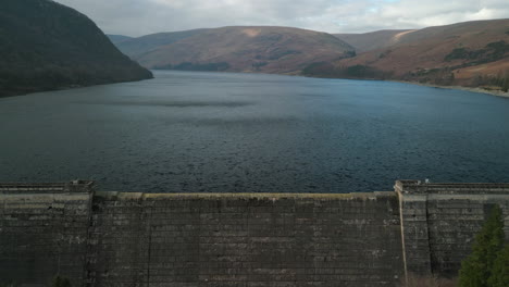Flying-towards-and-over-reservoir-dam-wall-to-windswept-lake-of-Haweswater-English-Lake-District-UK