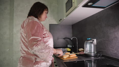 caregiver wearing pink robe prepares multiple sandwiches with cheese and meat slices on kitchen counter near electric kettle, apples, water bottles, and cutting board during early morning routine at home