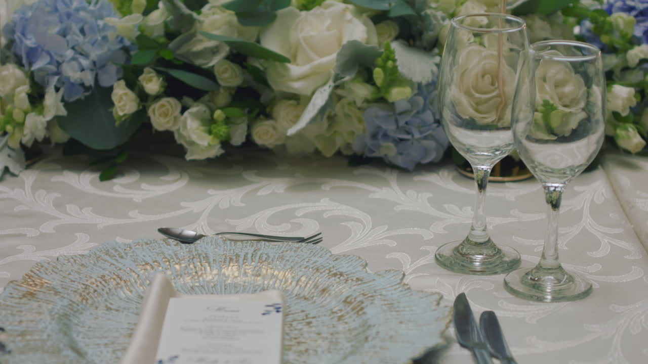 A detailed clip of an elegantly decorated wedding table. It features stemware, a white tablecloth, plates, cutlery, cloth napkins, a delicate floral arrangement, and a personalized invitation.