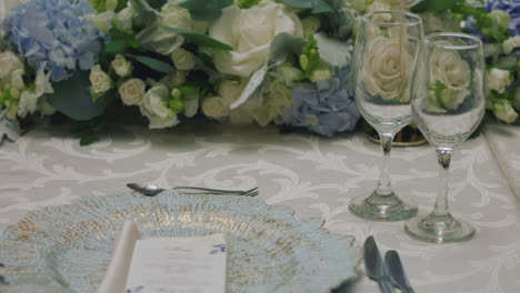 a detailed clip of an elegantly decorated wedding table. it features stemware, a white tablecloth, plates, cutlery, cloth napkins, a delicate floral arrangement, and a personalized invitation.