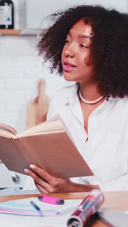 young african american woman student reads book sits at table in kitchen