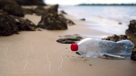 plastic bottle sits motionless amongst rocks on small sandy beach, water in background