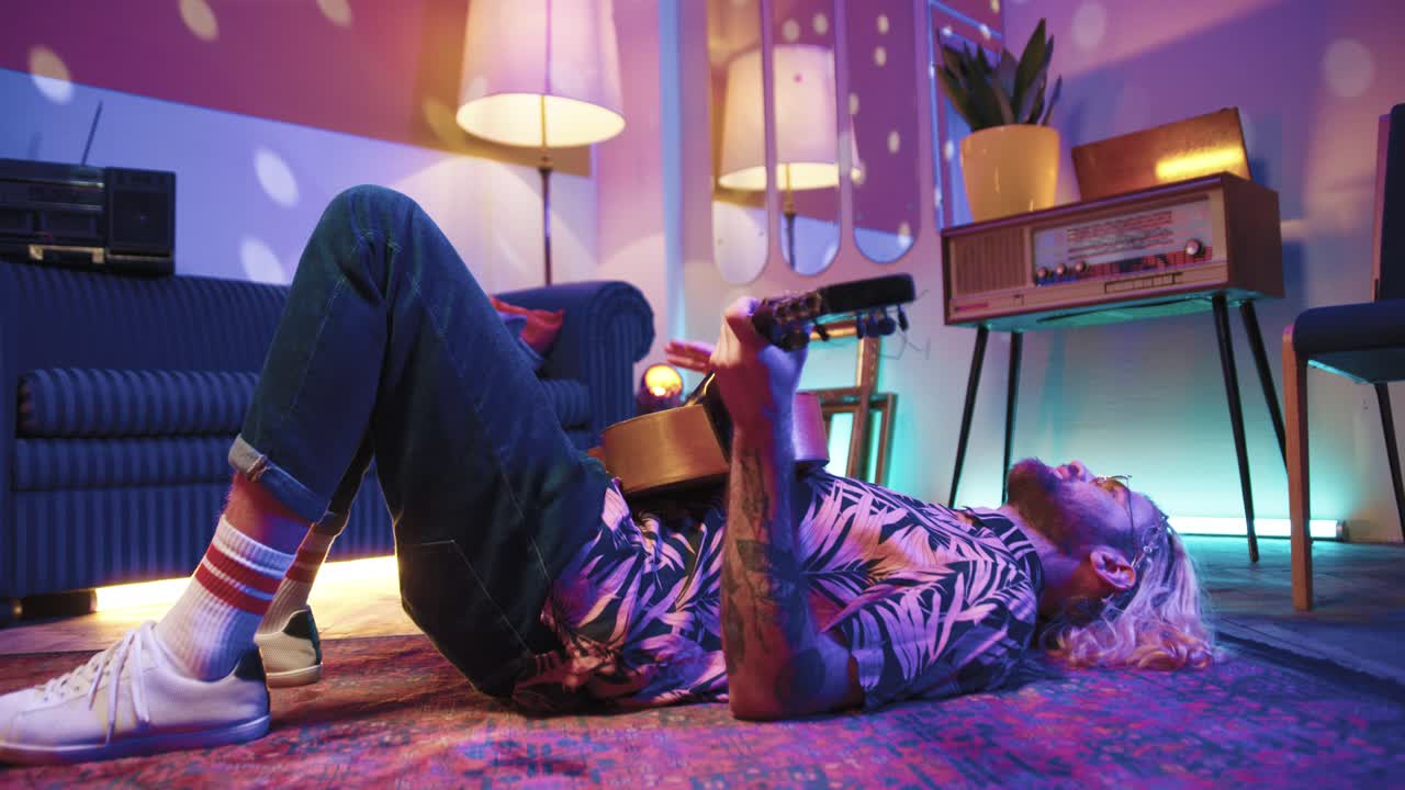 긴 머리를 바닥에 놓고 집에서 복고풍 파티에서 기타를 연주하는 세련된 백인 젊은이