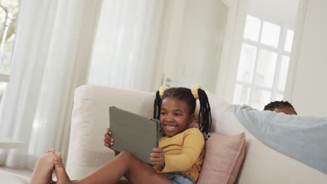 happy african american sister and brother using tablet on sofa at home, slow motion