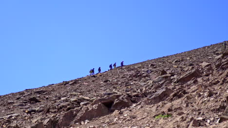 A-group-of-people-climbing-up-on-a-stony-mountain-ridge-in-the-distance,-against-clear-blue-sky-as-a-background