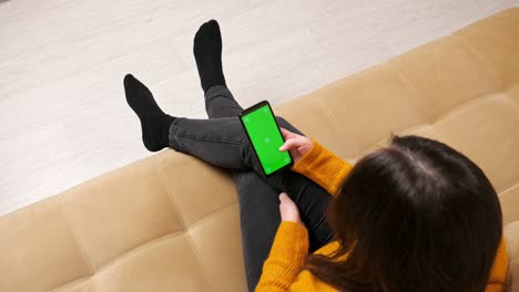 brunette in jeans sits on sofa with green screen smartphone
