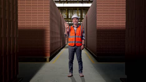 full body of asian male engineer with safety helmet smiling and showing okay gesture to the camera while standing with red brick packed in stacks are stored